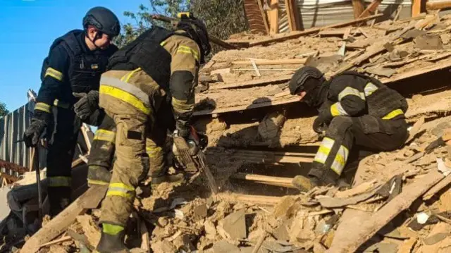 Emergency service workers on the roof of a building. The roof is destroyed with holes in it and debris all over it. Two men crouch and one sits looking down through a hole