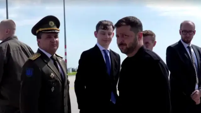 Zelensky looks side on at the camera. He wears black. There is a line of men, three in suits and one in an army uniform. They look on at Zelensky.