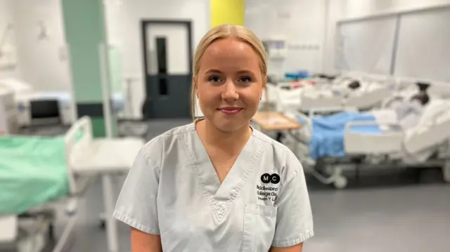 Freya is standing in front of a mock health ward wearing Middlesbrough College branded light grey scrubs.