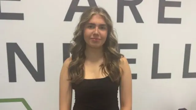 She is wearing a black vest top and has brown hair streaked with blonde in loose curls. behind her is writing on the wall, the only word of which is fully in the picture is the word are.