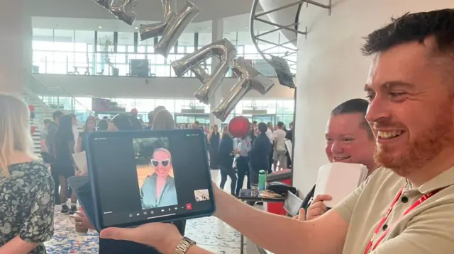A person holding an ipad and smiling. There's someone on a video chat on the ipad also smiling while wearing heart shaped sunglasses and a blue robe