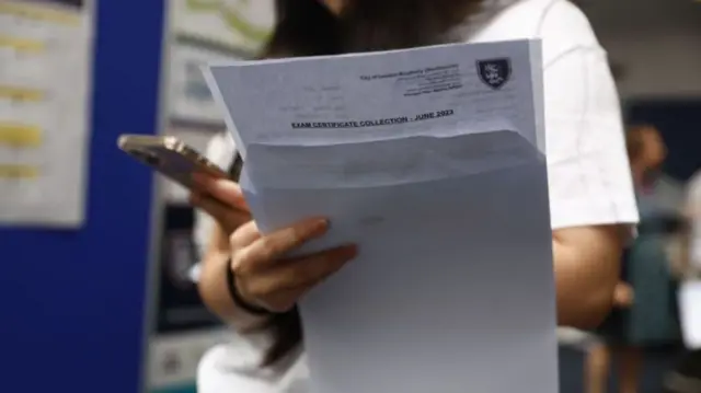a student hold their a-level results in their hand. with the other hand they are holding a phone.