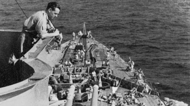 A man looks over the edge of a deck on a ship with men working below, in black and white