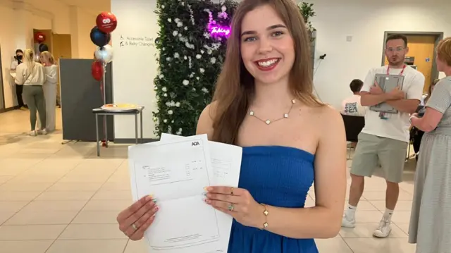 A girl smiling holding her results papers in a reception area of a school. People mill about behind her and there's some balloons to mark the occasion