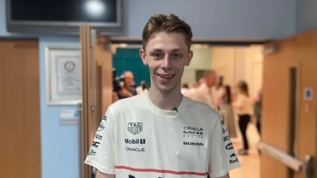 Harry smiles at the camera standing in the doorway of a school hall wearing an F1 t-shirt