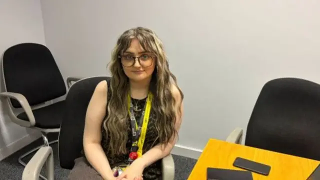 Kelsey is sat in a chair in an office with two phones on the desk and a black laptop. She's wearing smart trousers and a black vest top. She's also got yellow and black lanyards. Kelsey's wearing big frame glasses. Her hair is brown, wavey with streaks of blonde.