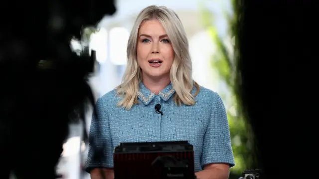 Karoline Leavitt, in a light blue blouse, speaks with a mic attached to her lapel