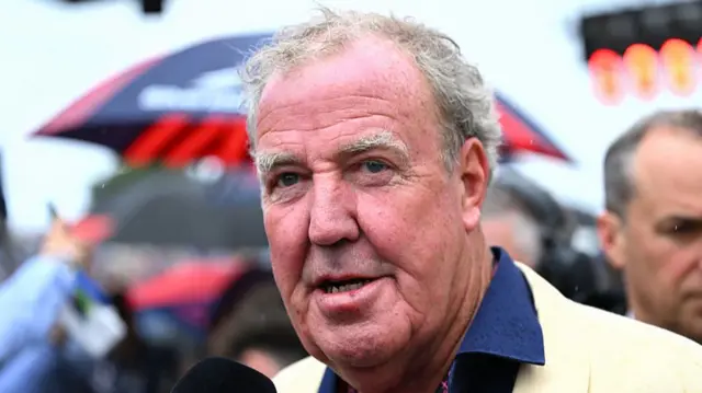 he is standing outdoors and there is a partial mic in front of him. In the background there is a blurry black and red umbrella and lots of people