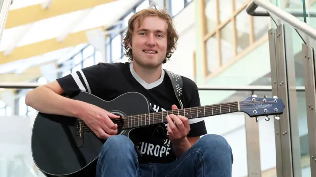 Dylan Griffiths wearing a black tshirt and smiling as he strums a black guitar. He appears to be sitting at the top of some stairs and in the background is a glass barrier