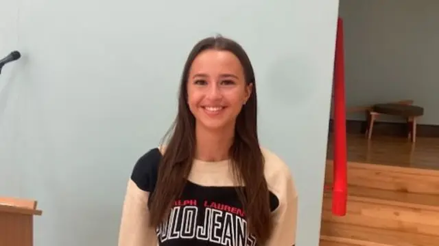 A brown haired girl smiling at the camera stood in front of a wooden set of steps and a wall. Next to her is a podium with two small microphones behind it.