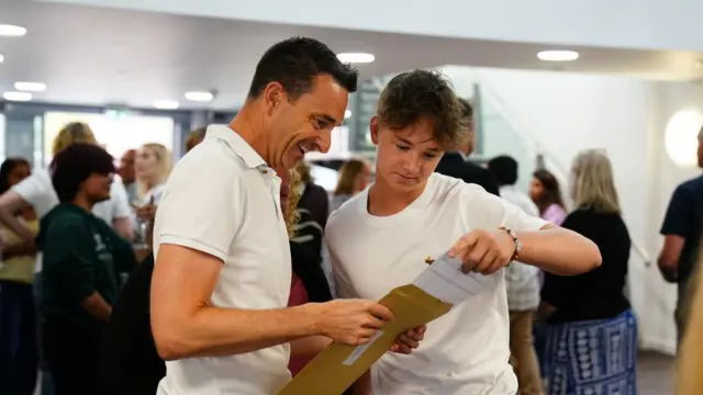 Two men, one older and one a student, look at a piece of paper they pull out of a brown envelope. One smiles broadly, the other slightly. The room is packed behind them