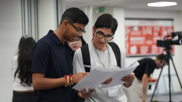 A boy and girl look together at their results. There's more people blurred in the background and a large tv camera