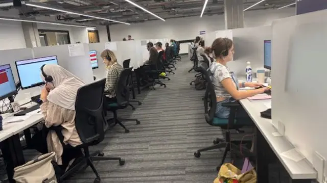 Two long rows of people sitting at desks with headphones on typing onto computers