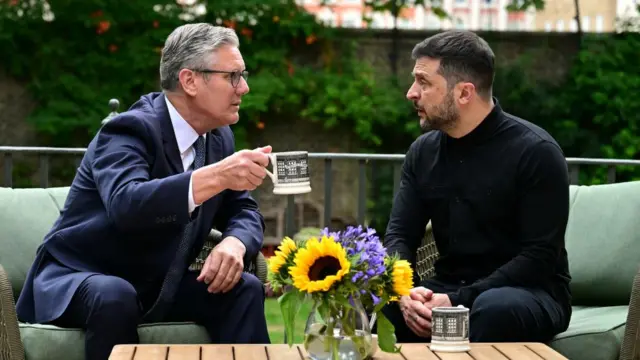 Starmer holds a mug, wearing a suit, and looks at Zelensky. They are both seated in the Downing Street garden.