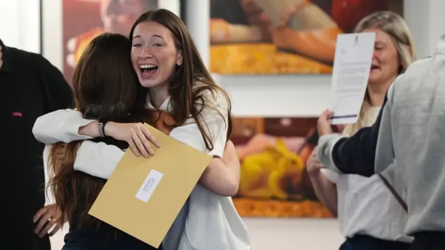 A brunette girl is hugging another girl while holding a brown A4 envelope. In the background on the right, there is a hand holding up a sheet of paper that is obscuring a woman's face
