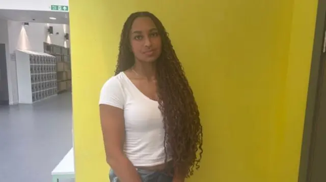 Headshot of Aisha who stands in front of a yellow wall with her arms in front of her and hands together