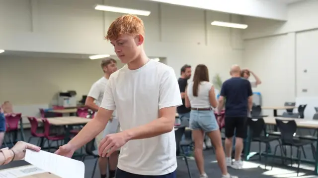 A boy takes a piece of paper from an outstretched hand. He stands in a classroom with people stood behind him
