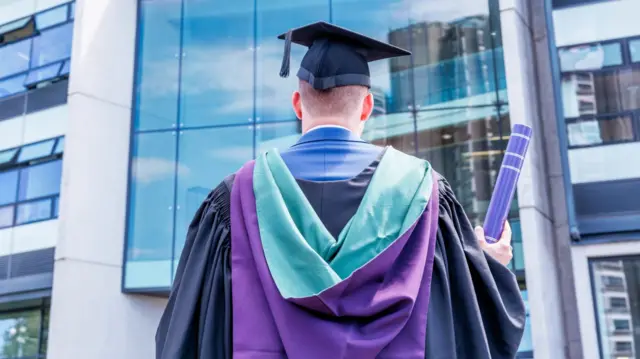 A man pictured from behind as he look on at a large modern building with large windows. He wears a graduation gown with a purple and blue hood, and holds a purple tube