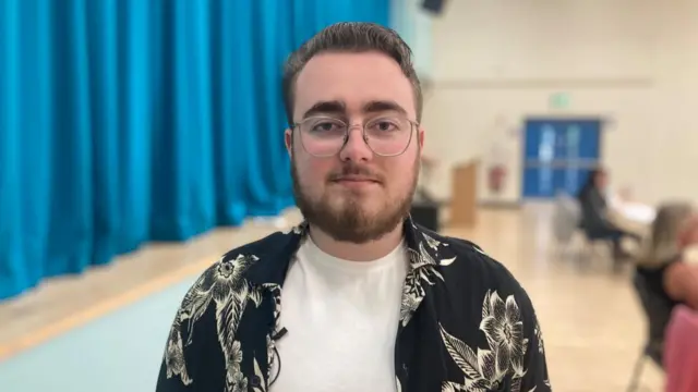 Max looking into the camera as he stands in front of a stage which has blue curtains pulled shut