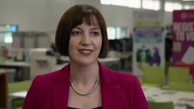 Bridget Phillipson smiling. She is wearing a magenta blazer and a silver necklace. In the background there are empty desks and chairs out of focus.