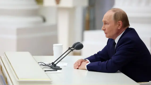 Putin, wearing a navy suit at a large white desk with gold edges in a grand-looking and white room, speaks into microphones during a meeting with members of Russia's leadership