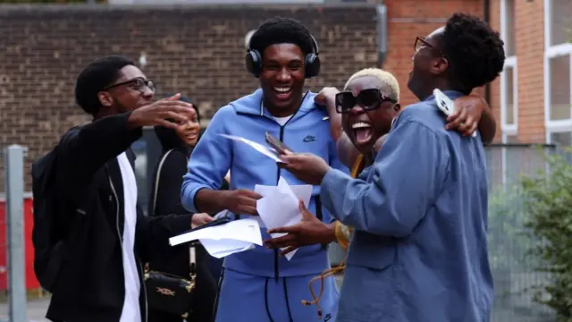 A group of four friends open their exam results and another classmate looks on in the background. They are smiling and and have their arms around one another. The student on the far left wears thick rimmed glasses and a black hoodie over a white t-shirt, and is facing the student to his right, who is wearing headphones and a blue tracksuit. Second from the right, another student with short blonde hair and sunglasses smiles towards the camera. Furthest on the right, a student in a blue shirt wearing glasses looks away from the camera towards the rest of the group