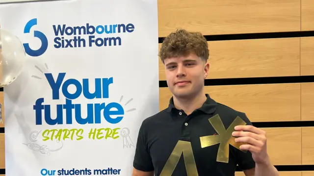 Sam holding up a gold star cutout and and A cutout in front of a sign that says Your Future starts here. He smiles