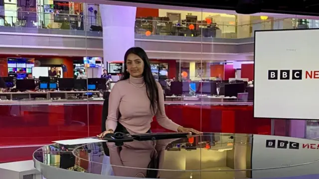 Annabelle sits at a large round table in front of a busy newsroom full of computer screens. There's a tv behind her where you can see the words BBC News