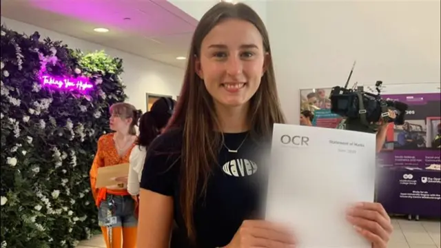 Grace smiling at the camera holding her results paper, which is blurred out. There's people standing in the background and a tv camera too.