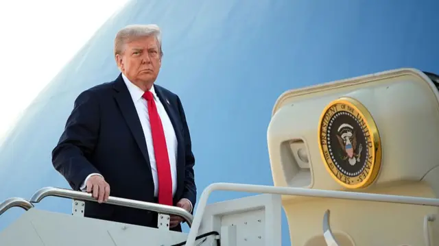 Donald Trump in a black suit, white shirt and red tie prepares to board Air Force One, his right hand is holding on to the metal railing of the stairs to the plane as he looks towards the tarmac