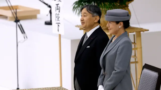 Both are standing. Naruhito is wearing a black suit, white shirt and grey tie. Masako is wearing a formal grey suit and matching hat. She has pearl stud earrings and a string of pearls around her neck. In the background is a bamboo table with a large flowerpot, and a white stage