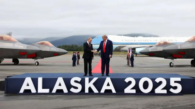 Vladimir Putin (L) shakes hands with Donald Trump (R) as they stand on a blue carpet covered platform, the words Alaska 2025 in white in front. Air Force One is visible on the tarmac in the background