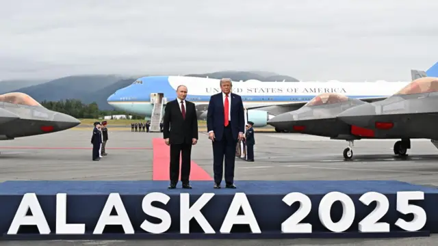 US President Donald Trump (right) and Russian President Vladimir Putin pose on a podium with the words: Alaska 2025 in white at the base. Air Force One and US fighter jets are behind them in the background.