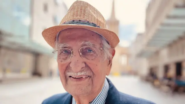 Captain Yavar Abbas, wearing a hat and blue coat, stands on a piazza