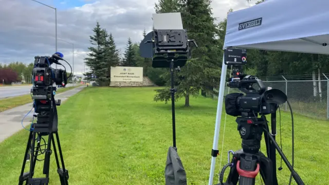 Cameras set on tripods and a media light pointed at a patch of grass outside the Elmendorf-Richardson military base in Anchorage