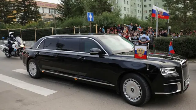 The Aurus Senate limousine carrying Russian President Vladimir Putin drives along a street in Ulaanbaatar, Mongolia. people are gathered on the side of the side and try to capture an image on their photos. The Russian and Mongolian flags are also in view.