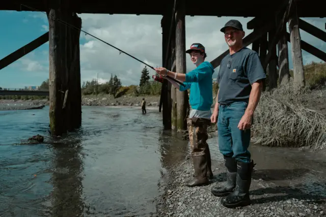 Two men wearing boots stand at side of river, one has fishing rod in his hands