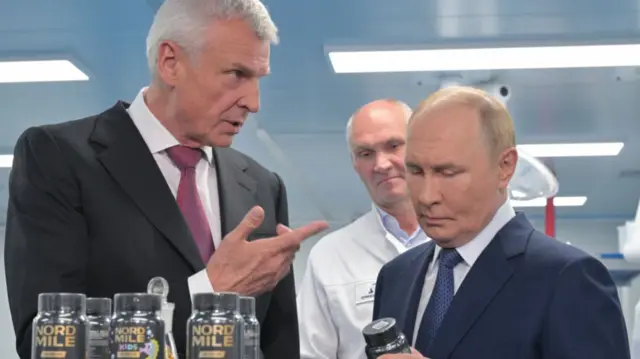 Vladimir Putin looks down at a black pill bottle as a man in dark suit to his right speaks to him. A man in a white lab coat stands behind them