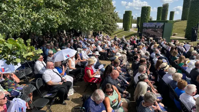 A crowd in the sunshine. Some have umbrellas or are wearing hats.