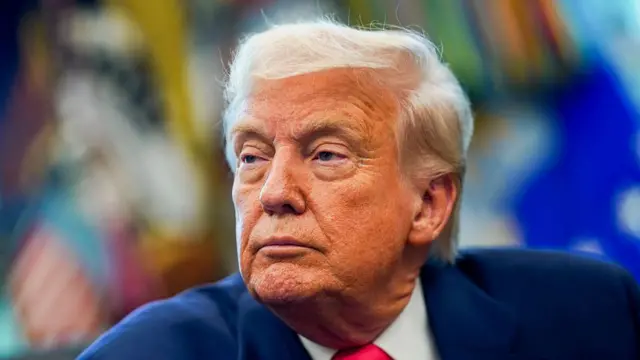 Trump wears blue suit, red tie and looks to the side in the Oval Office.