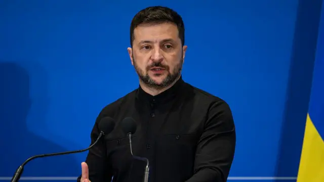 Ukrainian President Volodymyr Zelensky speaks into two thin mics on stage. He's in a black, long-sleeved top in front of a blue background, a Ukrainian flag to his left partially out of frame