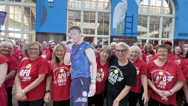 Members of the All For One choir, dressed in red, smile into the camera at the Paragon Interchange in Hull. They are standing with a cardboard cutout of Coldplay singer Chris Martin.