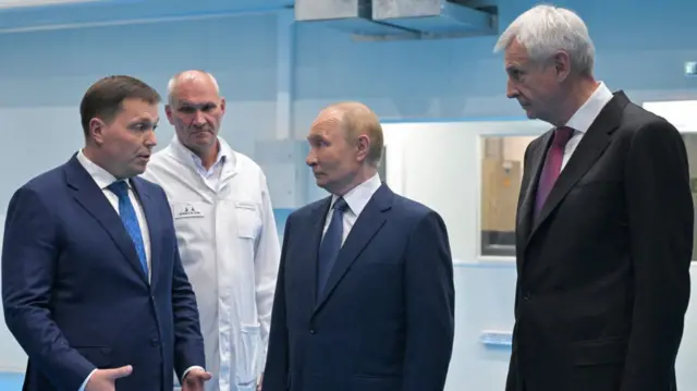 Vladimir Putin (C) in a dark suit stands between two men, one in a dark blue suit (L) speaking to him and a tall white-haired man in a black suit to his left. A man in a white lab coat listens in from the sidelines
