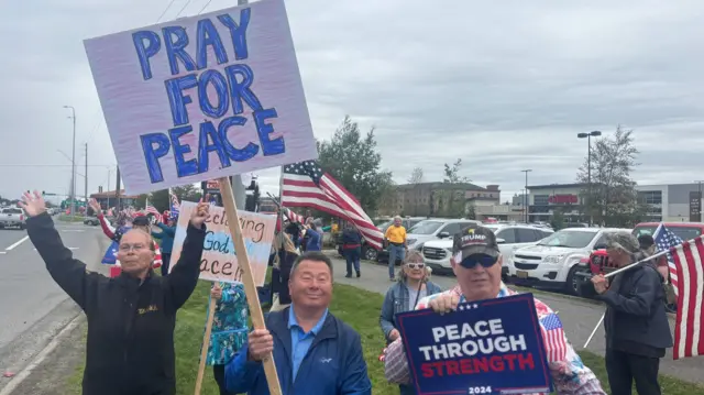 A woman with her hair pulled back in black clothing (L) holds up her arms. Next to her, a man in blue rain jacket and polo shirt (C) holds a sign reading Pray for Peace. A man in a pro-Trump cap and US flags jacket holds a sign reading Peace Through Strength in white and red ink over a blue background. Behind them other Trump supporters rally on a sidewalk next to a parking lot