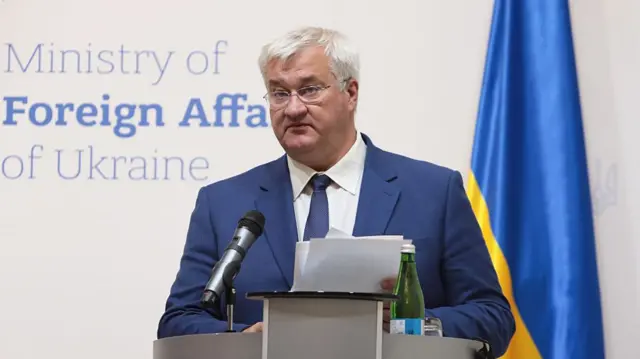 Ukrainian foreign minister Andrii Sybiha delivers a speech wearing a blue suit, matching tie and white shirt. Behind him, to the left is a Ukrainian flag leaning on a white wall, the phrase Ministry of Foreign Affairs of Ukraine written on it