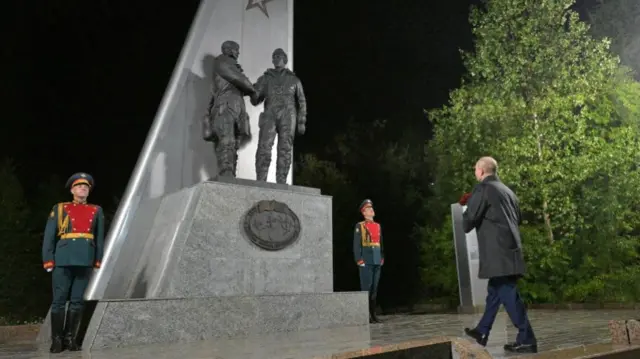 Russian-US World War Two memorial of Soviet and US pilots shaking hands. A Russian soldier is standing on either side of it as Putin walks towards it from the right. His back is to the camera. It is night.