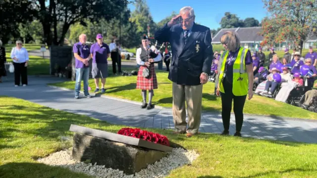 A veteran salutes in front of a memorial, which has had a poppy wreath laid on it