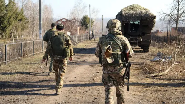 Russian soldiers walk along a street in a part of the Kursk region. In front of them is a patrol vehicle.