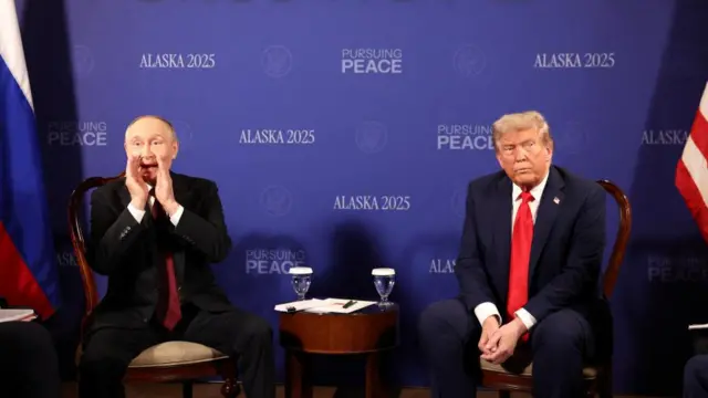 Putin and Trump sat next to each other inside, in front of a backdrop that says "Pursuing Peace" and "Alaska 2025".