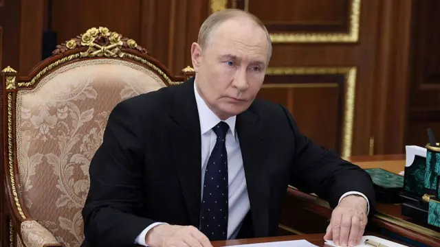 Vladimir Putin in Black suit, shirt and tie sits down on elaborate fabric chair inside a room with wood-covered walls
